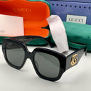 Black Gucci sunglasses with gold logo on a white surface, with Gucci packaging in the background.